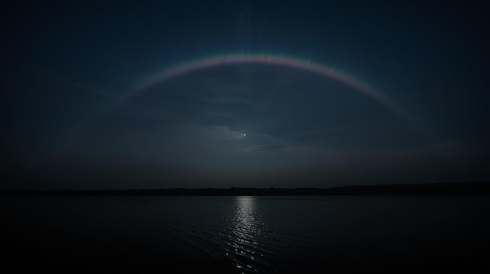 “Lunar rainbow appearing at night over a lake with moonlight reflection.”
